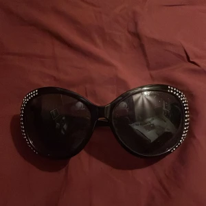 Svarta solglasögon med strassdetaljer - Snygga svarta solglasögon med stora bågar och glittrande strassdetaljer längs kanterna och skalmarna. Perfekta för att ge en glamorös touch till din outfit. Passar bra för både soliga dagar och festliga tillfällen.