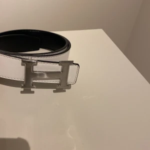 Svart och vitt bälte med silverfärgat spänne - Snyggt bälte i svart och vitt med ett stort, silverfärgat H-format spänne. Perfekt för att ge en stilren touch till din outfit. Bältet är vändbart vilket ger dig två olika looks i ett. Varan har inte använts