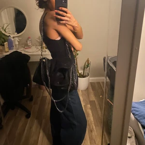 Svart axelväska med kedjedetaljer - Snygg svart axelväska med kedjedetaljer. Väskan har en glittrig yta och en stilren design som passar perfekt för olika tillfällen. Den har en praktisk storlek och en kedja som axelrem för en trendig look.