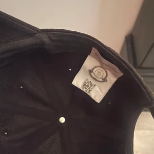 Svart keps från Moncler - Snygg svart keps från Moncler med broderad logga framtill. Justerbar rem med röd, vit och blå detalj baktill för perfekt passform. Perfekt för en stilren look.