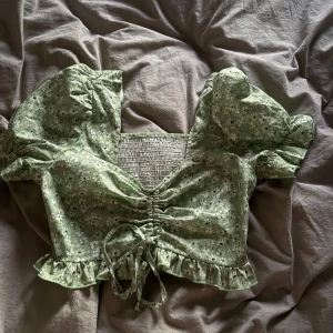 Grön blommig topp med volangdetaljer - Söt grön blommig topp med korta puffärmar och volangdetaljer. Toppen har en smockad rygg och knytning framtill för en justerbar passform. Perfekt för en somrig look!