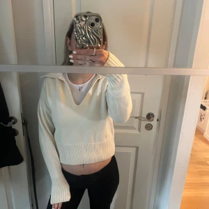 Vit stickad tröja med krage - Säljer en stilren vit stickad tröja med krage och v-ringning. Tröjan är croppad och har långa ärmar med ribbade muddar. Perfekt för en trendig look.