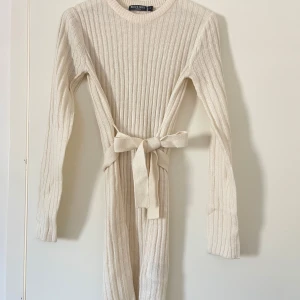 Beige ribbad klänning med knytband - Säljer en stilren beige ribbad klänning. Klänningen har långa ärmar och ett knytband i midjan för en snygg passform. Perfekt för både vardag och festliga tillfällen.