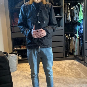 Svart vindjacka från Moncler - Säljer en stilren svart vindjacka från Moncler med dragkedja och en broderad logotyp på ärmen. Jackan har långa ärmar och en hög krage för extra skydd mot vinden. Perfekt för blåsiga dagar! Den är i ett jättefint skick, ingen låda eller något följer med för att den vid detta tillfälle inte är äkta