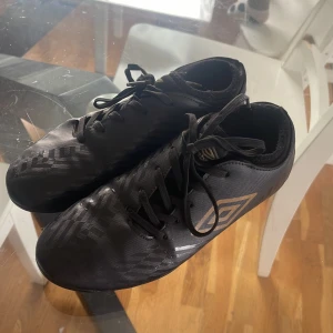 Svarta fotbollsskor från Umbro - Säljer ett par svarta fotbollsskor från Umbro med snörning och rund tå. Skorna har en diskret mönstring och är utrustade med dobbar för bra grepp på planen. Perfekta för fotbollsträning eller match.