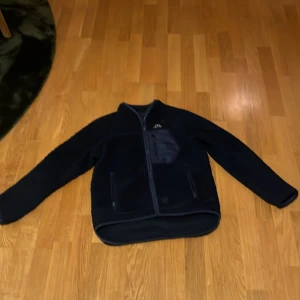 Mörkblå fleecejacka från Kappa - Säljer en svart fleecejacka från Kappa med dragkedja och två fickor framtill. Jackan har ett broderat Kappa-märke på bröstet och är perfekt för kyligare dagar. Den är långärmad och har en mjuk och varm känsla.