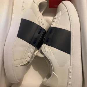 Valentino Garavani sneakers - Snygga vita sneakers från Valentino Garavani med en bred mörkblå rem över ovansidan. Skorna har en stilren design med rund tå och snörning. Perfekta för en trendig look.