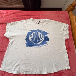 Vit t-shirt med blått tryck från Junk - Säljer en vit t-shirt från Junk med ett coolt blått tryck av en snäcka på framsidan. T-shirten har en avslappnad passform och korta ärmar, perfekt för en casual look. Materialet är mjukt och bekvämt.