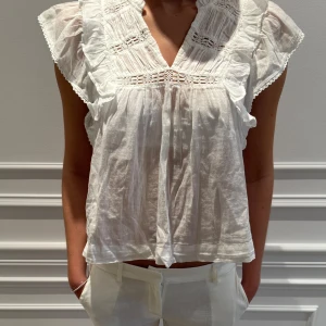 Vit blus från Neo Noir - Säljer en luftig vit blus från Neo Noir med volangdetaljer och spets. Blusen har en v-ringad design och korta ärmar med volang. Perfekt för en somrig look! Använd fåtal gånger.