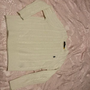 Vit stickad tröja från Ralph Lauren - Säljer en stilren vit stickad tröja från Ralph Lauren med det klassiska broderade logotypen på bröstet. Tröjan har en kabelstickad design och långa ärmar, perfekt för kyligare dagar. Passar både till jeans och kjol för en avslappnad look.