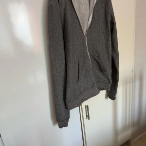 H&M  - Säljer en grå hoodie med dragkedja och fickor framtill. Perfekt för en avslappnad stil. Den har långa ärmar och en mjuk insida för extra komfort.