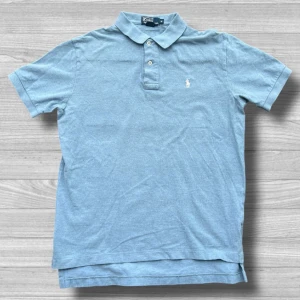 Ralph Lauren piké - Säljer en klassisk blå pikétröja från Polo Ralph Lauren med broderad logga på bröstet. Tröjan har korta ärmar och knappar vid kragen. Perfekt för en stilren look!