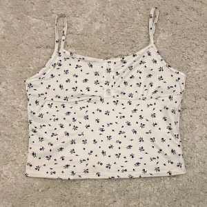 Vit blommig linnetopp  - Söt vit linnetopp från Shein med blått blommönster. Toppen har smala axelband och knappar framtill för en extra detalj. Perfekt för varma dagar!