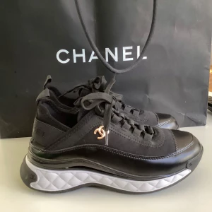 Svarta sneakers från Chanel - Snygga svarta sneakers från Chanel med vit sula och klassisk logga på sidan. Skorna har snörning och en bekväm, vadderad sula för extra komfort. Perfekta för en stilren look.
