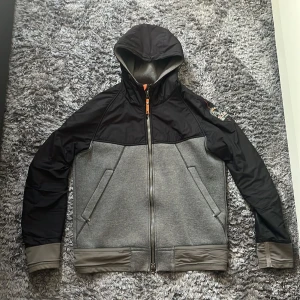 Grå och svart limited edition jacka från Parajumpers - Säljer en snygg grå och svart limited edition jacka från Parajumpers med huva och dragkedja. Jackan har en sportig look med kontrasterande färger och praktiska fickor framtill. Perfekt för en aktiv livsstil.