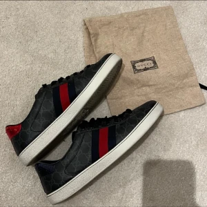 Gucci ace skor - Snygga svarta sneakers från Gucci med det ikoniska GG-mönstret. Skorna har röda och blå ränder på sidorna och en röd detalj på hälen. De är tillverkade i skinn och har snörning framtill. Perfekta för en stilren look.