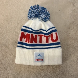 Minttu mösaa  - Säljer en vit mössa med blå och röda ränder samt en stor tofs på toppen. Mössan har texten 'MINTTU' i rött och en etikett framtill. Perfekt för kalla dagar! Hör av dig vid funderingar och pris kan diskutera vid snabb affär mvh Malte.202