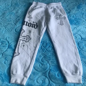 Vita mjukisbyxor med korsdetaljer - Snygga vita mjukisbyxor med tryck av kors och text i svart. Byxorna har elastisk midja och muddar vid bensluten för en bekväm passform. Perfekta för en avslappnad stil. Storlek s passar mer som M