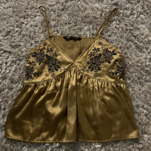 Fest topp från Zara - Snygg guldigt satinlinne från Zara med smala axelband och v-ringning. Toppen har vackra svarta broderade blommor på bröstet som ger en elegant touch. Perfekt för en stilfull look.