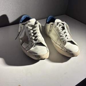 Golden goose skor - Snygga vita sneakers från Golden Goose med en ikonisk stjärndetalj på sidan. Skorna har en blå hälflik och snörning framtill. Perfekta för en avslappnad stil med en touch av lyx. Kan gå ner i pris vid snabba affärer 😇. Skriv om du har någon fråga. 
