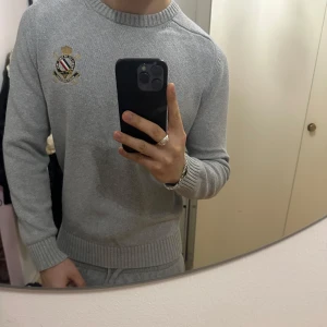 Grå stickad tröja från Morris Gents - Säljer en stilren grå tröja från Morris Gents med broderad logotyp på bröstet. Tröjan har en ribbad rund hals och långa ärmar, perfekt för en klassisk look. Passar bra till både jeans och chinos. 