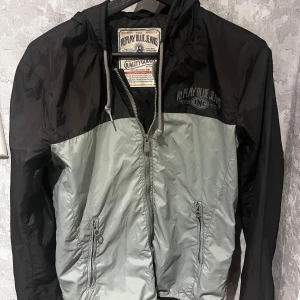 Svart och grå vindjacka från Replay - Snygg vindjacka från Replay i svart och grått. Jackan har en dragkedja framtill och justerbar huva med snörning. Perfekt för blåsiga dagar med sin lätta och funktionella design.