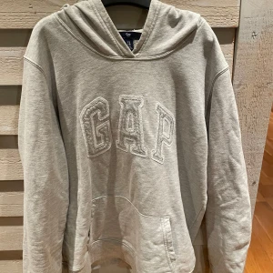 Grå hoodie från GAP - Säljer en klassisk grå hoodie från GAP med stor logga framtill. Tröjan har en bekväm passform och en praktisk känguruficka. Perfekt för en avslappnad stil. En fläck men man tänker inte på den!