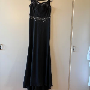 Svart långklänning med paljetter - Elegant svart långklänning med glittrande paljetter på överdelen och midjan. Klänningen har smala axelband och en figursydd passform som ger en stilren siluett. Perfekt för en glamorös kväll.