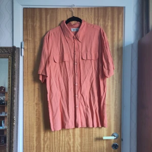 Korallfärgad skjorta från Blu di Blu - Mer orange än rosa! Snygg,  ostruken, korallfärgad skjorta från Blu di Blu med korta ärmar och knappar framtill. Perfekt för en avslappnad stil med två bröstfickor som ger en cool touch. Skjortan har en klassisk krage och är tillverkad i ett lätt material.