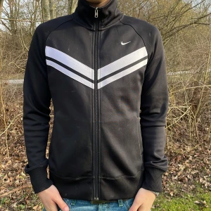 Svart träningsjacka från Nike - Säljer en svart träningsjacka från Nike med vita detaljer på bröstet. Jackan har en dragkedja framtill och långa ärmar. Perfekt för sportiga aktiviteter eller en avslappnad stil. Den har en hög krage och ribbade muddar för extra komfort.