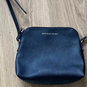 Svart axelväska från Victoria's Secret - Snygg svart axelväska från Victoria's Secret med en elegant design. Väskan har en justerbar axelrem och en dragkedja upptill. Insidan är fodrad i en ljusrosa nyans, vilket ger en fin kontrast. Perfekt för att bära dina nödvändigheter med stil.