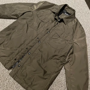 Olivgrön jacka från Massimo Dutti - Snygg olivgrön jacka från Massimo Dutti med quiltad insida. Jackan har knappar framtill och två bröstfickor. Perfekt för höst och vår med sin stilrena design och praktiska detaljer.