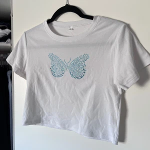 Vit croppad t-shirt med fjärilsmotiv - Säljer en vit croppad t-shirt med ett snyggt blått fjärilsmotiv i strass på framsidan. T-shirten har korta ärmar och är perfekt för en avslappnad stil. Passar bra till jeans eller kjol för en trendig look 🩵 Liten i storlek, passar bra till S/M. Använd en handfull gånger. Önskar du veta exakta mått? Tveka inte att höra av dig!