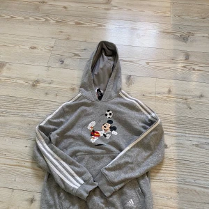 Grå hoodie med Musse Pigg från Adidas - Säljer en grå hoodie från Adidas med Musse Pigg som spelar fotboll på framsidan. Tröjan har de klassiska tre ränderna längs ärmarna och en bekväm huva. Perfekt för den sportiga looken!