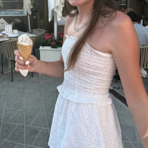 Vit axelbandslös klänning - Söt vit axelbandslös klänning med smockdetaljer och volangkant. Perfekt för varma sommardagar. Klänningen har en luftig och bekväm passform.