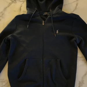 Svart huvtröja från Polo Ralph Lauren - Säljer en stilren svart huvtröja från Polo Ralph Lauren. Tröjan har dragkedja framtill och en broderad logotyp på bröstet. Den är utrustad med en huva och snörning för justering. Perfekt för en avslappnad look.