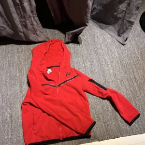 Röd huvtröja från Nike - Säljer en röd huvtröja från Nike med dragkedja och svarta detaljer. Tröjan har långa ärmar och en praktisk huva. Perfekt för en sportig look eller en avslappnad dag. 🏃‍♂️