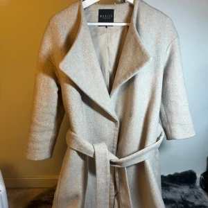 Beige kappa - Snygg beige kappa med ett stilrent och elegant snitt. Den har en bred krage och ett bälte i midjan för en fin passform. Perfekt för kyligare dagar.