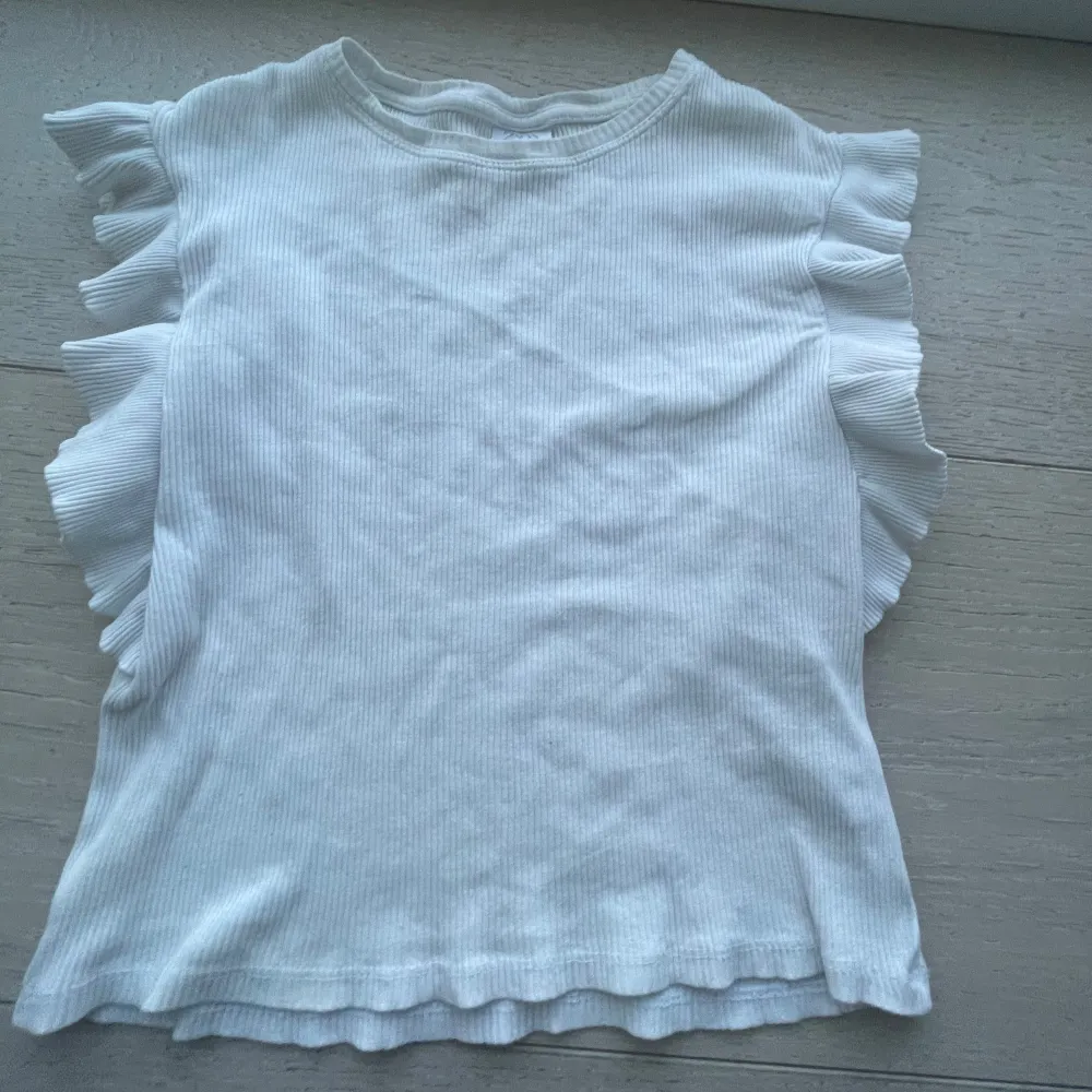 Säljer en söt vit ribbad topp från Zara med volangdetaljer längs ärmarna. Perfekt för en stilren och bekväm look. Passar bra till både jeans och kjol. 🌟. Yläosat.