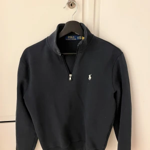 Mörkblå tröja från Polo Ralph Lauren - Säljer en stilren mörkblå tröja från Polo Ralph Lauren med dragkedja och klassisk logga på bröstet. Perfekt för en avslappnad och snygg look. Tröjan har långa ärmar och en hög krage.