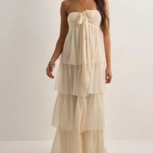 Créme färgad axelbandslös klänning - Elegant créme färgad axelbandslös klänning med volanger och knytband i nacken. Perfekt för sommarens festligheter med sin långa, flödande design.