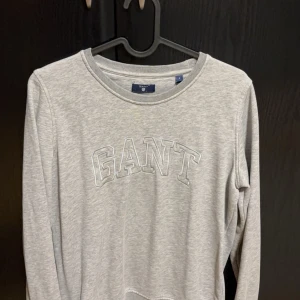 Grå långärmad tröja från Gant - Säljer en stilren grå långärmad tröja från Gant. Tröjan har en rund halsringning och Gant-loggan broderad på framsidan. Perfekt för en avslappnad och bekväm stil. Passar bra till jeans eller chinos.