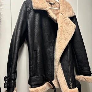 Svart skinnjacka med beige pälsfoder - Snygg svart skinnjacka med mjukt beige pälsfoder. Jackan har en asymmetrisk dragkedja och bältdetaljer vid ärmslut och midja. Perfekt för kyligare dagar med sin stilrena design och varma foder.