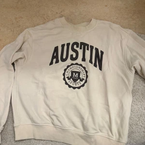 Beige sweatshirt från H&M Divided - Säljer en beige sweatshirt från H&M Divided med texten 'AUSTIN' och 'CHAMPIONSHIP SPORTS' tryckt i svart på framsidan. Tröjan har långa ärmar och ribbade muddar vid ärmslut och nederkant. Perfekt för en avslappnad stil.