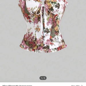 Blommig korsettopp med snörning - Snygg blommig korsettopp med snörning fram och bak. Toppen har volangdetaljer vid axlarna och nederkanten, vilket ger en romantisk touch. Perfekt för att lyfta vilken outfit som helst.