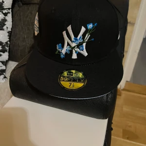 Svart keps från New Era - Snygg svart keps från New Era med broderad logga och blå blommor på framsidan. Kepsen är en 59FIFTY-modell med en klassisk passform. Perfekt för att ge din outfit en sportig touch.