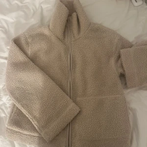 Beige teddyjacka med dragkedja - Mysig beige teddyjacka med dragkedja framtill. Perfekt för kyliga dagar med sin fluffiga och varma design. Jackan har långa ärmar och en hög krage för extra värme.