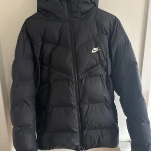 Svart dunjacka från Nike - Säljer en svart dunjacka från Nike med Storm-FIT teknologi. Jackan har en dragkedja framtill och är perfekt för kyliga dagar. Den har en klassisk design med Nike-logga på bröstet.🩷