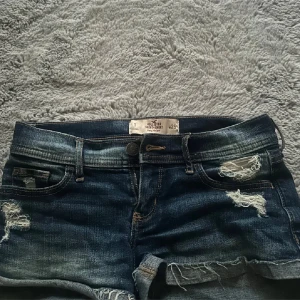 Mörkblå jeansshorts från Hollister - Snygga mörkblå jeansshorts från Hollister med slitna detaljer och låg midja. Perfekta för en avslappnad stil. De har en klassisk femficksdesign och knappgylf.