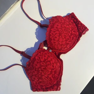 Röd spets-bh - Snygg röd bh med blommigt spetsmönster och justerbara axelband. Den har en liten dekorativ detalj framtill och hakknäppning bak. Perfekt för att ge en romantisk touch till din outfit.